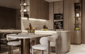 Une cuisine moderne avec des armoires en bois, un îlot incurvé avec des chaises blanches, des appareils électroménagers intégrés et des suspensions suspendues. Les plantes vertes ajoutent une touche de nature à cet appartement Dubaï.