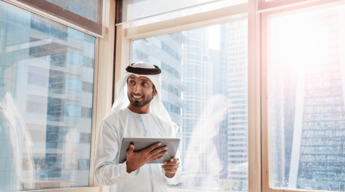 Emirati man in traditional dress smiling and holding a tablet, standing by a large window in his Dubai apartment with a view of the skyscrapers in the background.