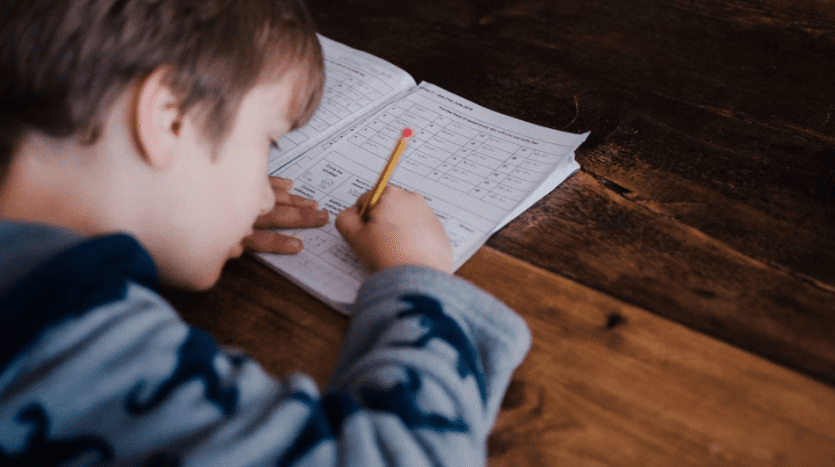 Un jeune enfant s'est concentré sur l'écriture dans un cahier avec un crayon, assis à une table en bois dans une villa à Dubaï. La vue montre le côté droit de l'enfant et le haut de sa tête.