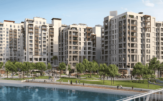Un complexe d'appartements moderne composé de plusieurs immeubles de grande hauteur surplombe un front de mer paisible. La zone comprend des pelouses verdoyantes, des palmiers et des sentiers pédestres, avec plusieurs personnes profitant de l'espace extérieur. Le ciel clair suggère une journée ensoleillée au bord de la lande.