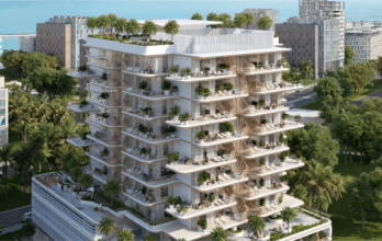Un immeuble d'appartements moderne à plusieurs étages entouré d'arbres et doté de grands balcons verdoyants se dresse fièrement près de Sunset Bay. La structure offre un aperçu de l'océan en arrière-plan, en harmonie avec les autres bâtiments voisins le long du front de mer tranquille.
