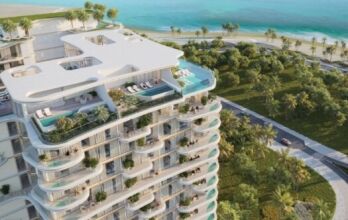 Beach Walk Grand 2 est un immeuble d'appartements moderne et luxueux en bord de mer, avec des balcons incurvés, des piscines sur le toit et une végétation luxuriante donnant sur une plage tropicale et un océan turquoise, entourés de palmiers et de jardins paysagers.