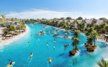 Le grand lagon bleu clair de Damac Islands 2 est entouré de villas modernes, de palmiers et de plages de sable, où les gens font du kayak, du paddleboard et se détendent sur des plates-formes flottantes sous un ciel d'un bleu éclatant.
