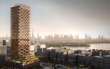Un grand gratte-ciel moderne à la façade unique en forme de grille, appelé The Symphony, se dresse parmi des immeubles de faible hauteur, surplombant une rivière et une ville lointaine sous un ciel nuageux.