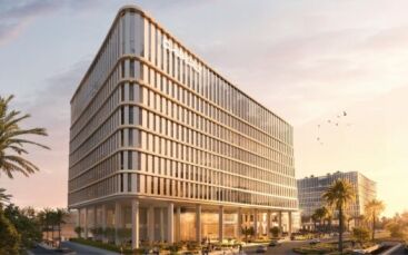 A modern multi-story office building with glass facades stands at sunset near Piazza Roma, surrounded by palm trees and landscaped walkways. People stroll outside while birds fly overhead. Another similar building is visible in the background.