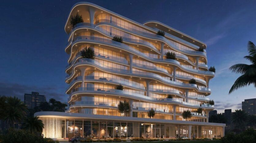 Un bâtiment Coralis moderne de plusieurs étages avec des balcons courbés en forme de vagues et des lumières brillantes au crépuscule, avec une végétation luxuriante sur les terrasses et entouré de palmiers dans un environnement urbain.