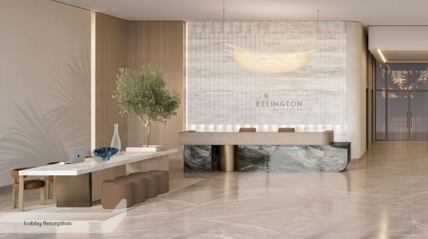 Le hall d'accueil moderne de l'Everly Place by Ellington se distingue par son sol en marbre, une longue table blanche accompagnée de tabourets marron, un arbre en pot et un comptoir d'accueil arborant le logo d'Ellington Properties. Un éclairage tamisé et des tons neutres viennent rehausser l'atmosphère accueillante.