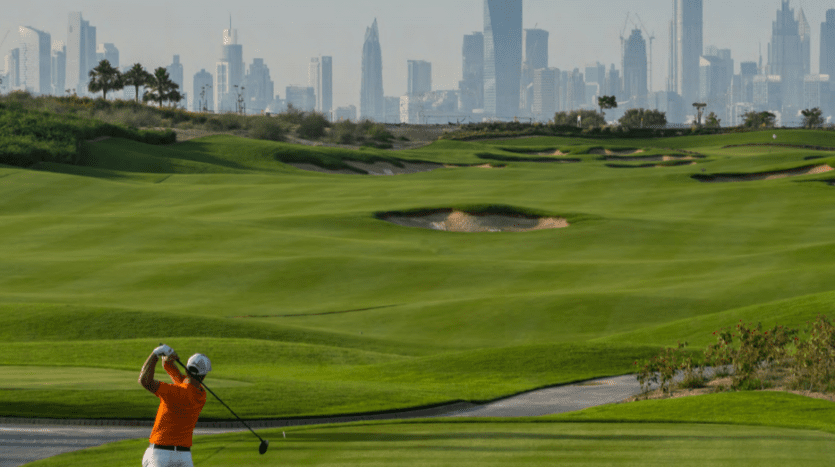 Un golfeur en chemise orange balance un club sur les greens luxuriants et vallonnés du parcours de golf de Dubai Hills avec l'horizon d'une ville, y compris d'imposants gratte-ciel, en arrière-plan.