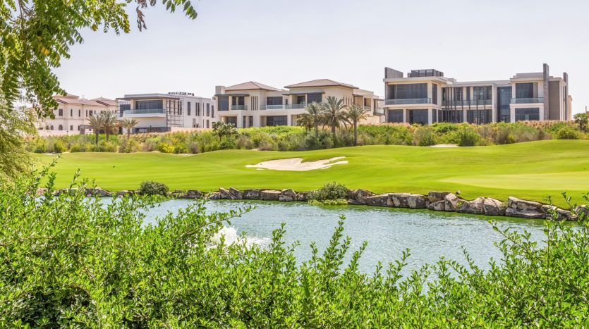 Un parcours de golf luxuriant avec un étang pittoresque au premier plan, entouré d'une verdure vibrante et présentant une rangée de maisons modernes et luxueuses en arrière-plan sous un ciel dégagé sur les collines de Dubaï.