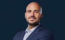 Une photo professionnelle d'un homme chauve souriant portant un costume, sur un fond bleu foncé, idéale pour les profils de l'immobilier Dubaï.