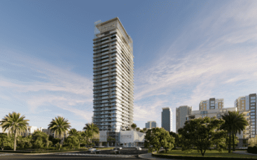 Le gratte-ciel moderne et de grande hauteur connu sous le nom de Skyhills 2 arbore une façade en verre saisissante, se dressant fièrement sur un ciel partiellement nuageux. Entouré de palmiers et d'une végétation luxuriante, il domine des bâtiments plus petits tandis que quelques voitures passent sur la route en face.