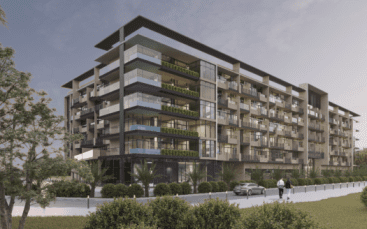 Un immeuble moderne de six étages avec des balcons en verre et de la verdure. Une personne marche sur le chemin et une voiture est garée à proximité. La scène se déroule sous un ciel nuageux et est entourée d'arbres et de buissons.