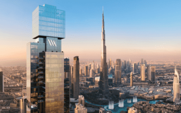 Un grand gratte-ciel moderne portant le logo "Waldorf Astoria" se dresse au premier plan, surplombant un paysage urbain composé du Burj Khalifa et des bâtiments environnants de Dubaï, sous un ciel clair au coucher du soleil.
