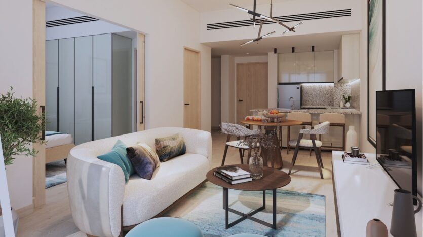 Salon et cuisine modernes à aire ouverte dans le Damac District de Dubaï, avec un canapé blanc, des coussins colorés, une table basse ronde en bois, une table de salle à manger, un décor minimaliste, et une chambre à coucher avec une armoire en verre visible à travers une porte coulissante.