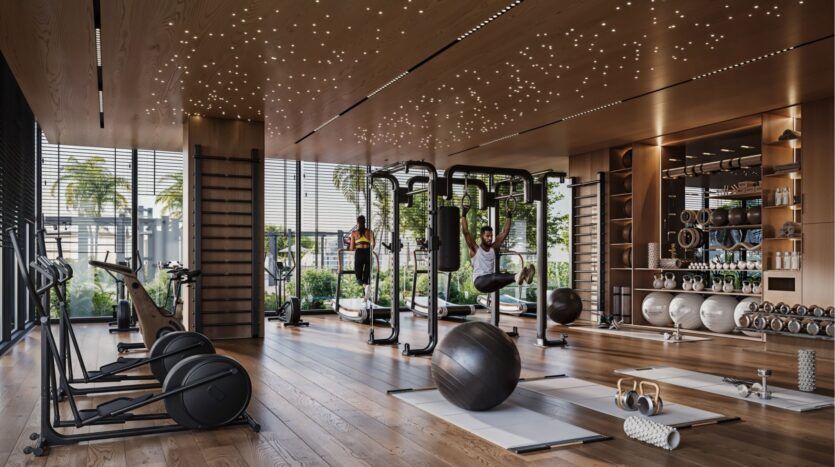 Salle de sport moderne dans le Damac District de Dubaï avec des sols en bois, de grandes fenêtres, des vélos d'exercice, des poids libres, des tapis de yoga et des équipements de remise en forme. Deux personnes s'entraînent tandis que la verdure brille à l'extérieur et que les plafonniers créent un effet de ciel étoilé.
