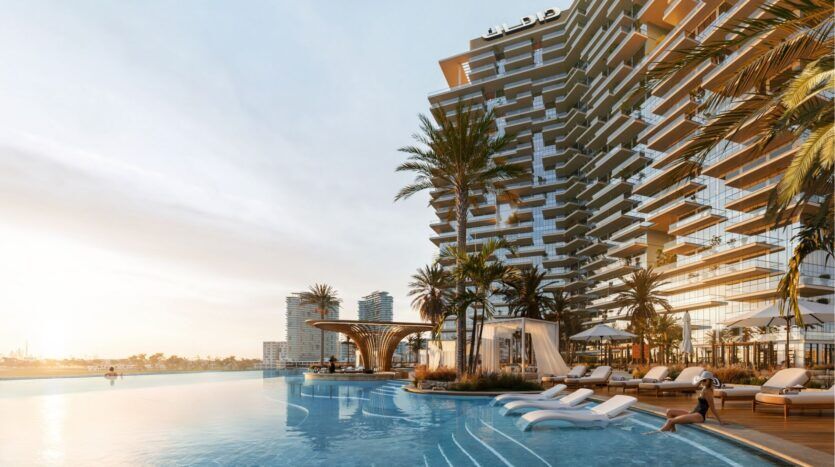 Un luxueux complexe situé au bord de l'eau dans le quartier Damac de Dubaï présente des terrasses modernes, des chaises longues au bord d'une piscine à débordement, des palmiers et des cabanas au coucher du soleil, avec les bâtiments de la ville visibles à l'arrière-plan.