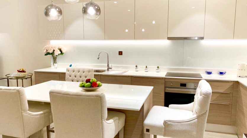 Une cuisine moderne avec des armoires blanches et en bois élégantes, un îlot blanc avec trois chaises rembourrées et des suspensions suspendues au-dessus. Le décor comprend des fleurs de Q Gardens Dubai et un bol de fruits.