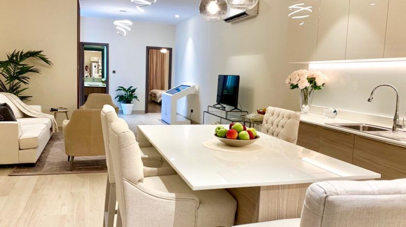 Cuisine moderne avec armoires blanches et coin repas comprenant une grande table blanche, des fleurs et un bol de fruits. Le plan ouvert débouche sur un salon avec des canapés et une télévision. Lumineux, chaleureux