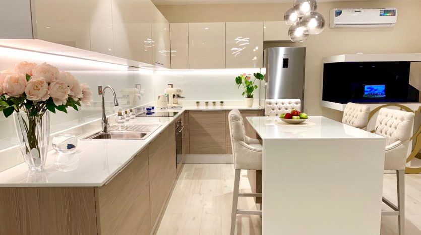 Intérieur de cuisine moderne aux tons neutres comportant un îlot central, des armoires en bois et équipée d'électroménagers haut de gamme. Une élégante salle à manger inspirée de Q Gardens Dubai, avec des sièges moelleux, est visible dans le