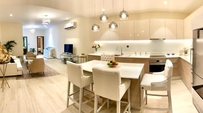 Intérieur de cuisine moderne avec armoires blanches, un long îlot avec tabourets de bar et un coin repas intégré. Un éclairage lumineux et des appareils élégants rehaussent l'aspect spacieux et épuré inspiré de l'esthétique de Q Gardens Dubai.