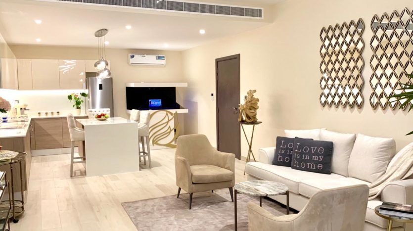 Un salon moderne et cosy comprenant un canapé blanc, deux chaises beiges et une télévision murale. Une cuisine ouverte avec un comptoir de bar et des tabourets est visible en arrière-plan. Le décor comprend de l'art mural