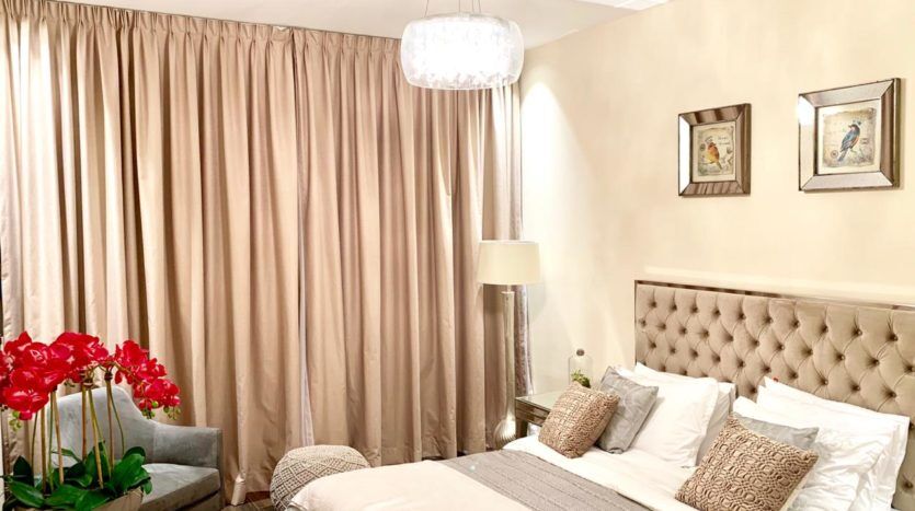 Une chambre confortable dotée d'une tête de lit capitonnée moelleuse, de rideaux beiges, d'un lustre élégant et d'un arrangement de fleurs rouge vif à côté du lit inspiré de Q Gardens Dubai.
