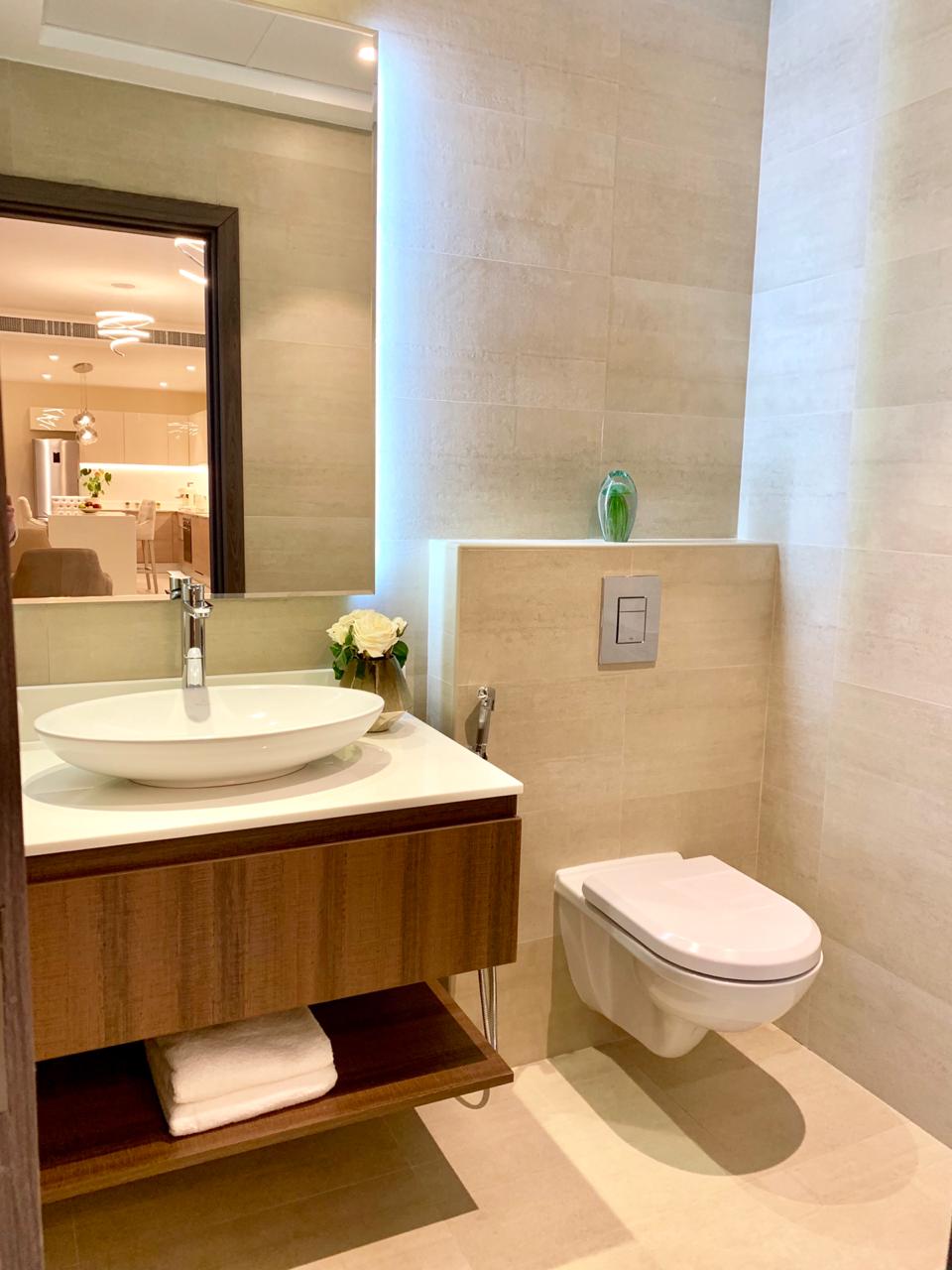 Salle de bain moderne aux murs beiges comportant une vasque ovale blanche sur un meuble vasque en bois, des toilettes à droite et un miroir visible en arrière-plan. Un éclairage doux améliore l'ambiance calme inspirée par Q Gardens Dubai