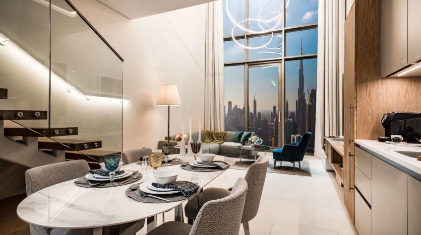 Une salle à manger et un salon modernes à SLS Dubaï avec une grande fenêtre donnant sur les toits de la ville, une table à manger pour quatre, des chaises élégantes et un luxueux canapé bleu.