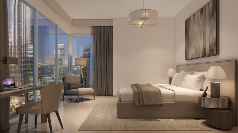Une chambre moderne avec un grand lit, un bureau et un fauteuil, dotée de baies vitrées donnant sur le paysage urbain de Dubaï la nuit. Tons neutres et décoration contemporaine.
