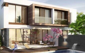 Une maison moderne de deux étages à Damac Hills avec de grandes fenêtres et des touches de bois, entourée d'une verdure luxuriante. Un arbre à fleurs roses fleurit au bord d’une piscine avec une personne se relaxant sur une chaise longue.