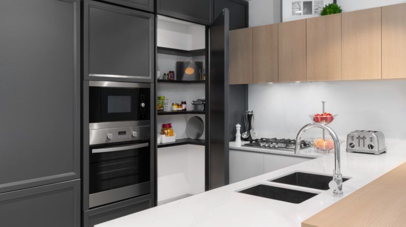 Cuisine moderne avec armoires noires et bois à Wilton Park, avec appareils électroménagers intégrés et comptoir blanc. Comprend un four, un four micro-ondes, une cuisinière et un bol de fruits