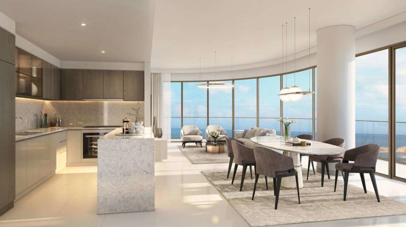 Intérieur d'appartement moderne et décloisonné comprenant une cuisine avec îlot en marbre, un coin repas avec une table blanche et des chaises grises et un coin salon avec des canapés, le tout donnant sur l'océan à travers des baies vitrées
