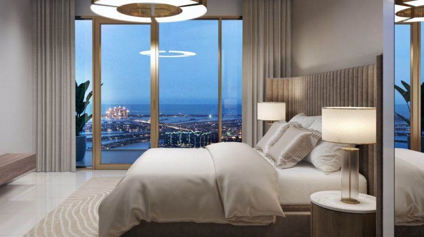 Une chambre luxueuse la nuit avec un grand lit, des lampes élégantes et des baies vitrées donnant sur le paysage urbain et l'océan bien éclairés du front de mer d'Elie Saab Emaar.
