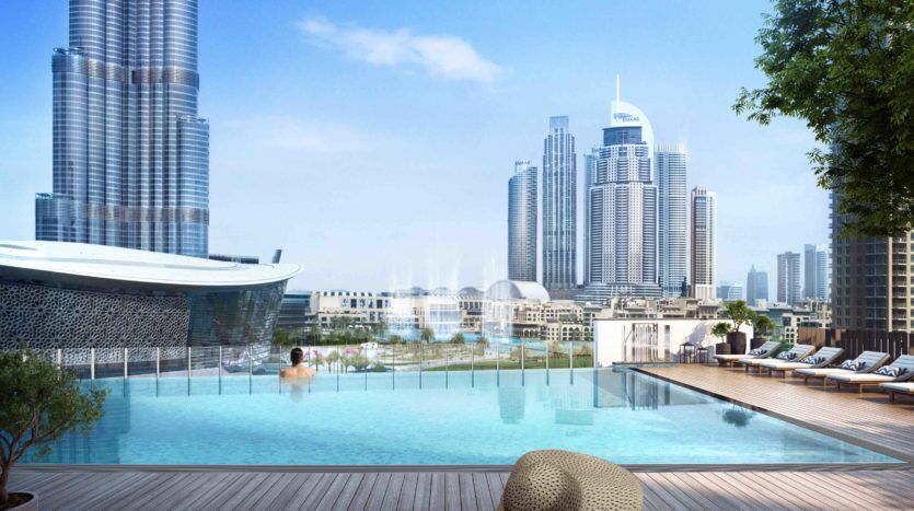 Une personne portant un chapeau est assise au bord d’une luxueuse piscine sur le toit surplombant un paysage urbain avec des gratte-ciel sous un ciel bleu clair. Des chaises longues blanches sont soigneusement disposées sur une terrasse en bois, mettant en valeur le véritable