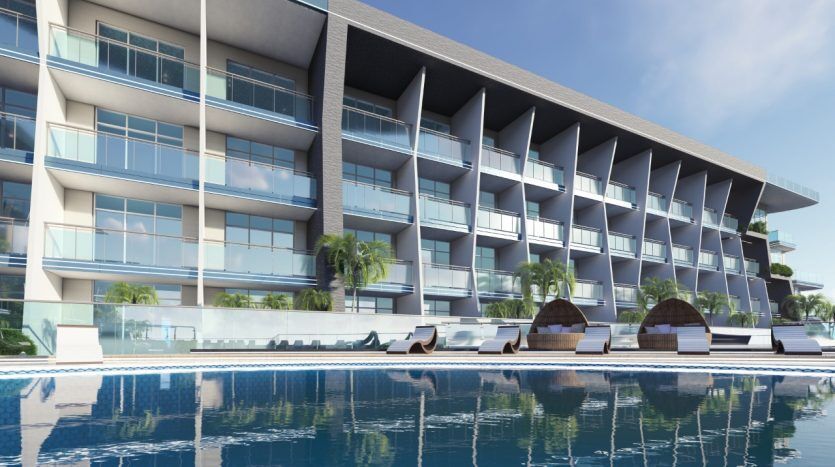 Hôtel moderne en bord de mer avec plusieurs balcons, fenêtres en verre réfléchissant et piscine bleu clair bordée de chaises longues et de cabanes sur Samanag Golf Avenue dans un cadre ensoleillé.