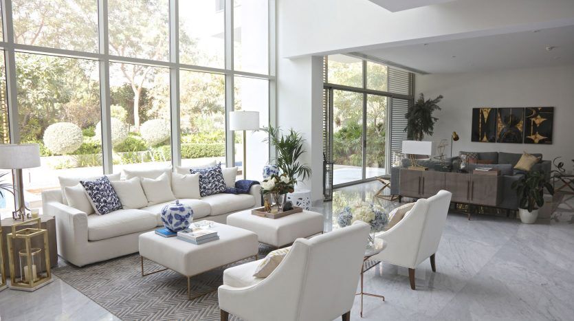Salon spacieux à la décoration moderne dans un appartement prestigieux de Dubaï, doté de grands canapés blancs, de sols en marbre et de baies vitrées donnant sur un jardin. Le décor comprend des accents bleus, des lampes et des éléments abstraits