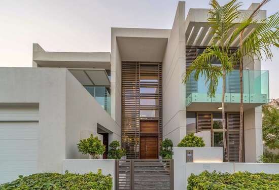 Villa moderne sur deux étages dans un quartier à l'architecture géométrique, dotée de grandes fenêtres, d'une porte en bois et d'un aménagement paysager subtil sous un ciel dégagé au crépuscule.