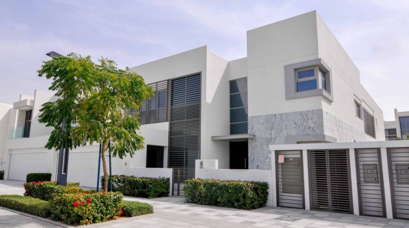 Villa moderne blanche sur deux étages dans un quartier à toit plat, comprenant un garage, une cour avant paysagée avec des fleurs et un petit arbre, sous un ciel dégagé.