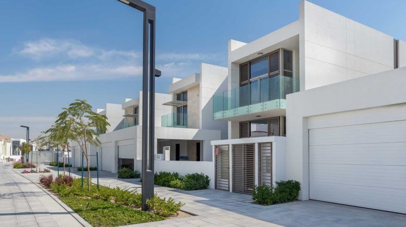 Une rangée de maisons blanches modernes aux lignes épurées, avec des balcons et de grands garages sous un ciel bleu clair, adjacentes à une rue soigneusement pavée avec de jeunes arbres à Dubaï.