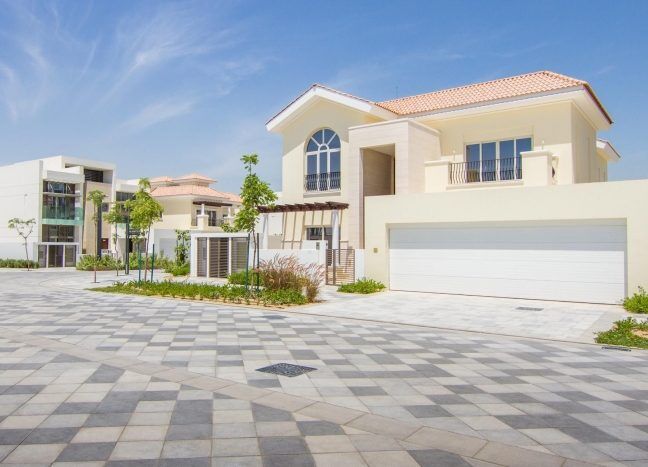 Un quartier de villas moderne avec de grandes maisons dotées de garages proéminents et de toits de tuiles sous un ciel bleu clair, avec un trottoir à motifs au premier plan.