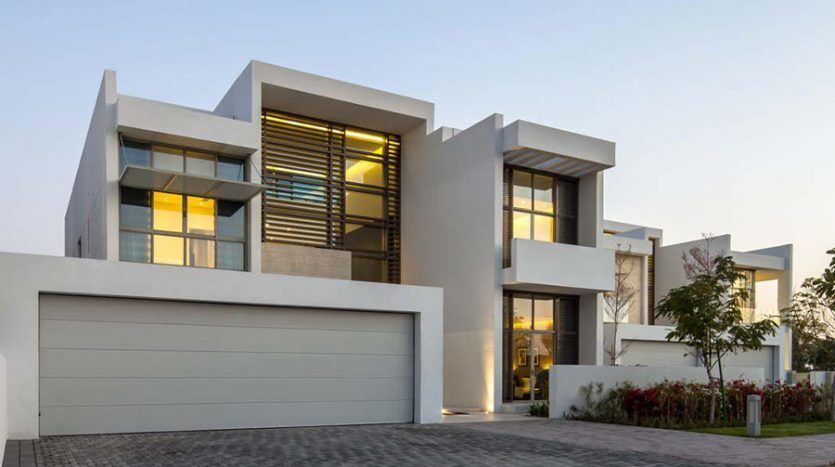 Maison moderne à deux étages avec un toit plat, de grandes fenêtres avec un éclairage intérieur chaleureux et une double porte de garage fermée, photographiée au crépuscule avec un ciel clair à Dubaï.