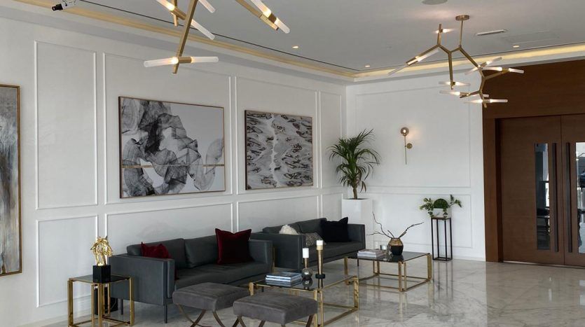 Lobby moderne et élégant avec sol en marbre blanc, grandes œuvres d'art abstraites sur murs lambrissés blancs, sièges élégants gris et noirs, tables basses géométriques dorées et lustres dorés distinctifs offrant une vue imprenable sur Dubaï.