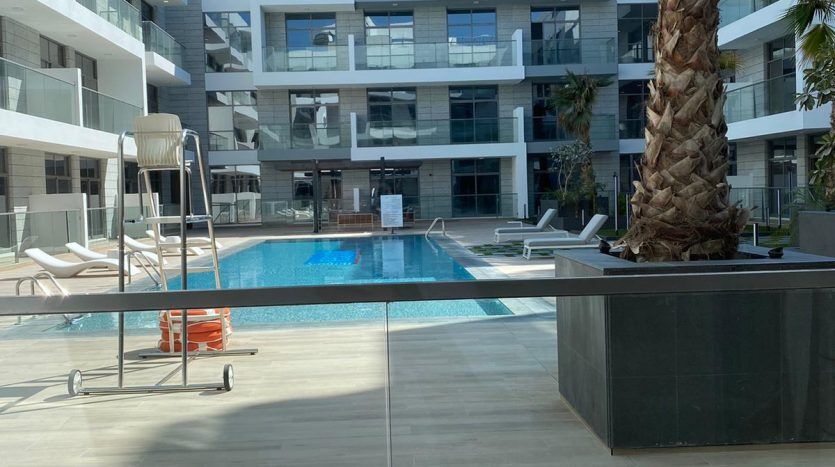 Un complexe d'appartements moderne à Dubaï avec une piscine bleu clair au centre, entourée de chaises longues et d'un grand palmier. Les bâtiments ont une façade blanche contemporaine avec des balcons offrant des vues privilégiées.