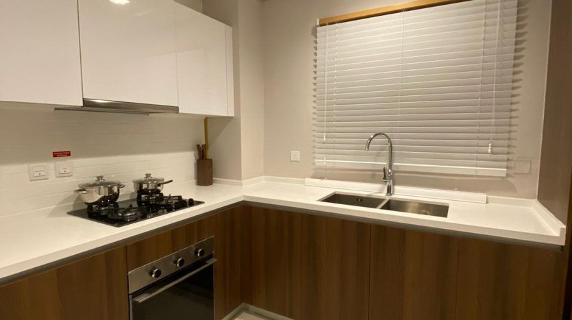 Intérieur de cuisine moderne avec comptoirs blancs, armoires en bois, cuisinière et four intégrés et fenêtre offrant une vue imprenable sur Dubaï au-dessus de l'évier.