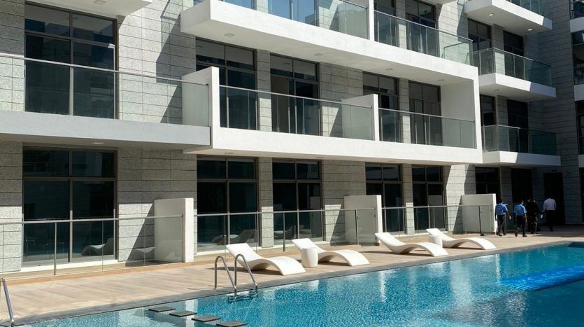 Un bâtiment moderne avec des balcons blancs donnant sur une piscine bleu clair entourée de chaises longues, sur fond de ciel dégagé, offrant une vue imprenable sur Dubaï.