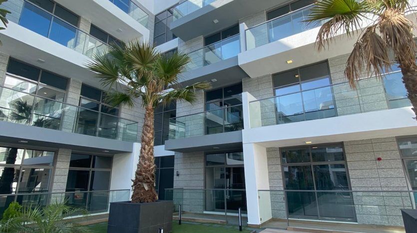Bâtiment urbain moderne avec balcons en verre, entouré de palmiers sur un patio carrelé sous un ciel bleu clair, offrant une vue imprenable sur Dubaï.