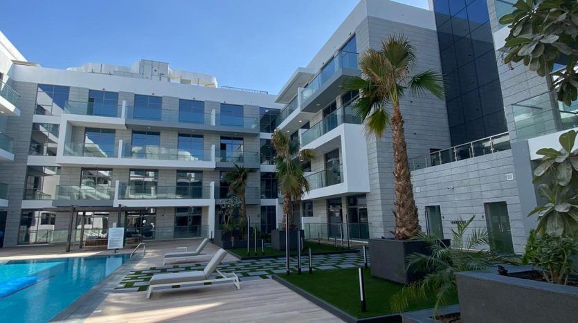 Bâtiment résidentiel moderne à Dubaï avec une piscine extérieure entourée de palmiers et de chaises longues, offrant une vue imprenable, un ciel bleu clair et une architecture géométrique.