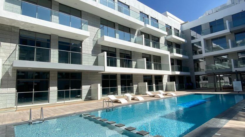 Un complexe d'appartements moderne à Dubaï avec une grande piscine dans la cour, entouré de bâtiments blancs à plusieurs étages sous un ciel bleu clair.