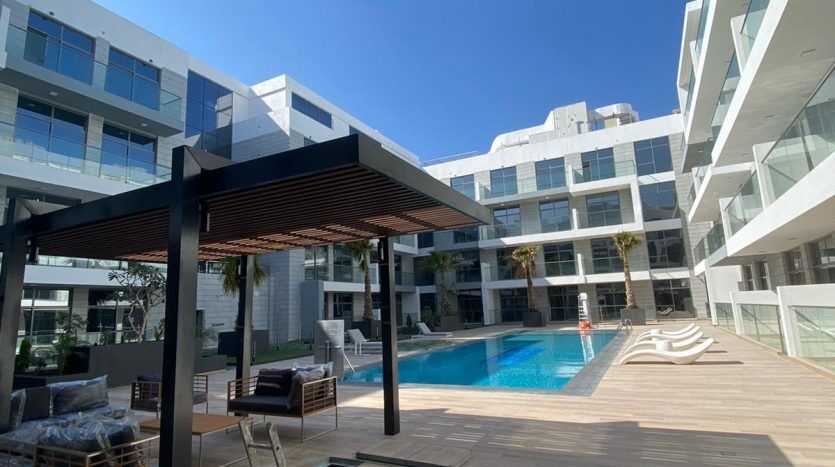 Un espace moderne au bord de la piscine dans un complexe d'appartements luxueux à Dubaï, doté d'un mobilier d'extérieur élégant sous une pergola, entouré de bâtiments contemporains à plusieurs étages par une journée ensoleillée.