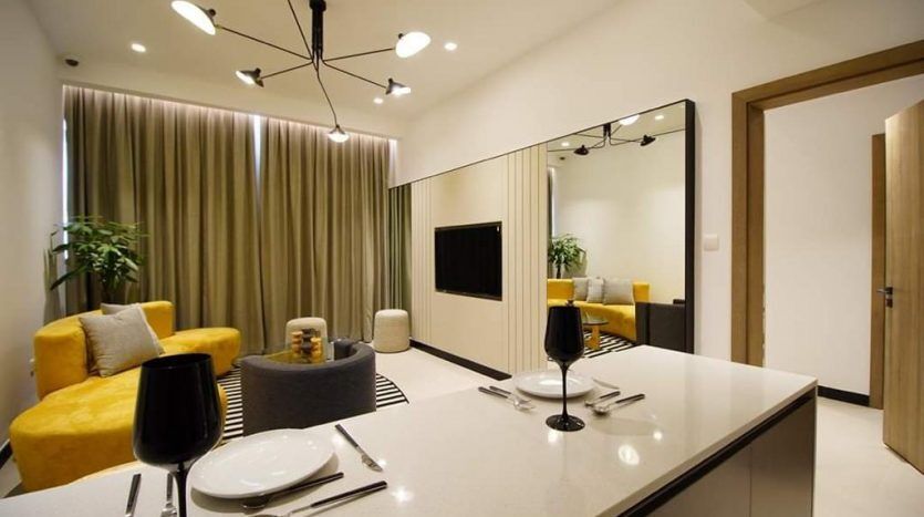 Salon moderne de l'Aria Résidence Dubai avec un canapé jaune, une table à manger en verre, un lustre unique et un mur en miroir reflétant une télévision et une autre partie de la pièce.